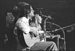 Deux personnes sont assises sur des chaises, jouant de la guitare et chantant dans des microphones, et d’autres personnes se trouvent derrière elles, dans l’ombre.