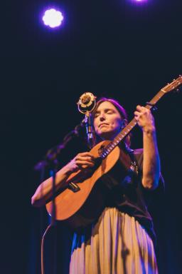 Une femme jouant de la guitare avec émotion sur une scène sombre. Elle se tient derrière un micro, les yeux fermés.