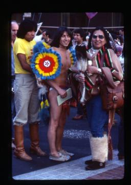 Une photo montrant 3 personnes au premier plan et une foule à l’arrière-plan. À droite, une personne portant des lunettes de soleil et des plumes dans les cheveux est vêtue d’un haut rayé, d’un jean et de mukluks en fourrure, tient un éventail en plumes d’aigle et sourit à l’appareil photo. La personne au centre ne porte que des mocassins, des sous-vêtements et quelques pièces d’un costume de pow-wow coloré et orné de plumes sur les épaules et le dos.