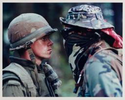 Un face-à-face tendu entre un soldat en uniforme portant un casque et une personne portant une veste de camouflage, un chapeau et un bandana couvrant son visage. 
