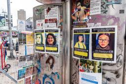 Un mur extérieur couvert de graffitis et d’affiches montrant des visages d’enfants et de jeunes avec du texte. En arrière-plan, un paysage urbain dans ce qui ressemble à une place de marché.