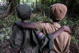 Deux enfants vus de dos, les bras passés sur les épaules l’un de l’autre. Celui de gauche porte un uniforme vert militaire avec un insigne du Sud-Soudan sur le bras droit et l’autre porte un uniforme beige. Tous deux portent un chapeau assorti à leur uniforme.