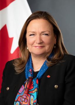 Photo de Marie-Geneviève Mounier, une femme blanche aux cheveux bruns mi-longs, portant un tailleur sombre, un foulard aux couleurs vives représentant une œuvre d’art autochtone et une épinglette à l’effigie de l’érable canadien.