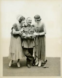 Photo en noir et blanc de deux hommes déguisés en femmes des années 1920. Les deux sourient de manière adorable et tiennent les bras de l’officier en uniforme qui se tient entre eux.