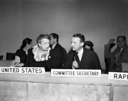 Une femme, Eleanor Roosevelt, est assise sur une estrade et écoute attentivement l’homme à sa gauche, John Peters Humphrey. Devant elle, une plaque porte l’inscription « United States ». Devant lui, une plaque indique « Secrétaire du comité » en anglais.