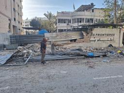 Un homme portant un chandail gris et un pantalon gris se tient devant le mur endommagé d’un bâtiment de Médecins sans frontières/Doctors Without Borders au milieu de débris et de décombres.