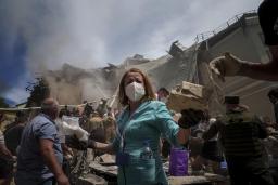 Une infirmière en blouse bleue portant un masque médical se tient au premier plan d’un hôpital détruit, entourée d’autres personnes civiles et militaires. De la fumée s’échappe de l’arrière près d’un bâtiment dont le toit s’est effondré.