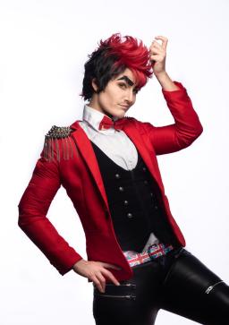 Un drag king vêtu d’une extravagante veste rouge à épaulettes, d’un gilet noir et d’un pantalon en cuir noir prend une pose et une expression coquines. Ses cheveux sombres sont coiffés d’une boucle rouge choquante et ses sourcils épais et collés sont pailletés.