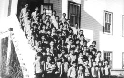 Sur cette photo d’archives en noir et blanc, des dizaines de jeunes élèves autochtones sont vêtues en uniforme de filles, c’est-à-dire en robe et en béret, et se tiennent sur les marches d’un grand escalier menant au bâtiment scolaire. Au premier rang du groupe se trouvent cinq religieuses en habit et deux prêtres en soutane.