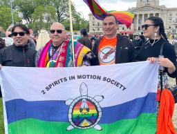 Lors d’une manifestation, quatre personnes autochtones tiennent un drapeau représentant une tortue, au centre de laquelle se trouvent des symboles bispirituels, dont deux plumes d’aigle réunies sur fond d’arc-en-ciel. Un drapeau arc-en-ciel flotte derrière les quatre personnes.