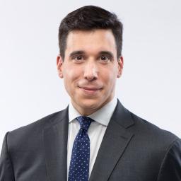 Un homme blanc aux cheveux foncés sourit légèrement à la caméra. Il porte un costume sombre avec une chemise blanc cassé et une cravate bleu foncé à petits pois blancs.