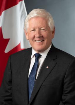 Un homme blanc aux cheveux gris se tient debout, souriant devant un drapeau canadien. Il porte un costume noir, une cravate bleue et une chemise blanche.