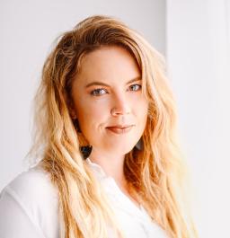 Une femme blanche aux longs cheveux blonds et portant un anneau dans le nez regarde la caméra. Elle porte une chemise blanche.