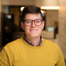 Une femme aux cheveux très courts et foncés regarde la caméra et sourit, la bouche fermée. Elle porte de grandes lunettes rondes à monture rouge et un chandail couleur moutarde par-dessus une chemise à carreaux à col. La pièce derrière elle est floue.