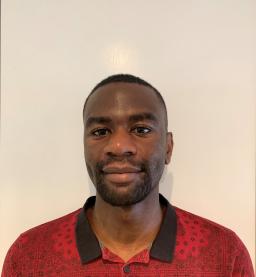 Une personne noire aux cheveux noirs très courts regarde la caméra avec un léger sourire. Elle porte une chemise rouge avec un motif imprimé représentant un dessin au trait noir et un col noir.