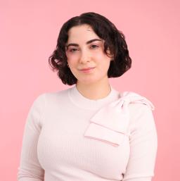 Une femme blanche aux cheveux bruns sourit à la caméra. Elle porte un gilet rose pâle avec un nœud rose sur l’épaule gauche.