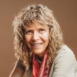 Une femme d’apparence blanche aux cheveux blonds bouclés sourit à la caméra. Elle porte une chemise beige et un foulard à motifs rouges.