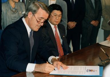 Brian Mulroney assis à une table, signant un document. M. Art Miki est assis à ses côtés et l’observe.