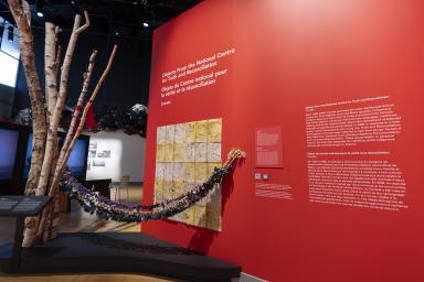 Des cordes auxquelles sont attachés des dizaines de sachets en tissu coloré sont suspendues entre un bouleau et un mur rouge.