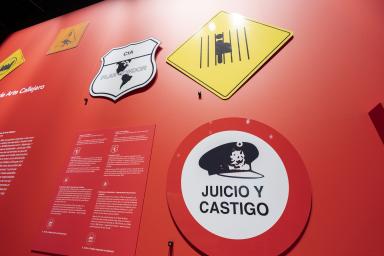 Des panneaux avec des éléments graphiques et des textes en espagnol sont accrochés à un mur rouge. Le plus grand panneau indique « Juicio y Castigo ».
