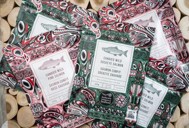 Des paquets avec des motifs colorés sur le thème des Haïdas sont empilés. Le texte en gros caractères sur les emballages indique «Saumon sockeye sauvage confit» ou «Saumon rose sauvage confit». Autre texte indique «Oncorhynchus nerka» , «60g», et, en anglais, «ready to eat», «sustainable», et «Authentic Indigenous Seafood».