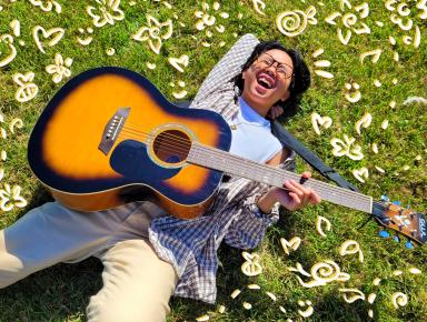 Kris Cahatol est en position allongée sur de l’herbe verte et rit. Des fleurs et des cœurs dessinés par ordinateur l’entourent. Kris tient une guitare sur sa poitrine.