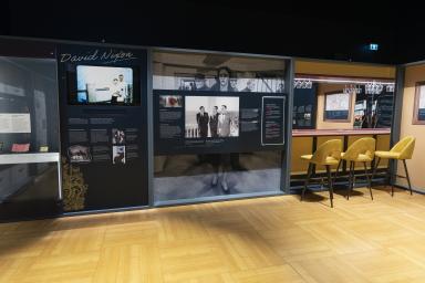 Une exposition muséale présente trois sections distinctes. Deux d’entre elles présentent des photos et du texte, tandis que la troisième est une reproduction d’un bar avec un comptoir et trois chaises jaunes.