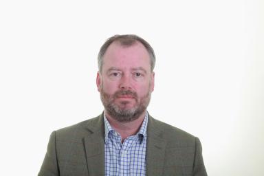 Un homme blanc aux cheveux grisonnants courts et à la barbe regarde la caméra d’un air impartial. Il porte un blazer gris et une chemise à carreaux bleus et blancs.
