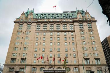 Un grand hôtel historique avec une façade en pierre beige, un toit en cuivre vert et des détails architecturaux ornés. Un drapeau canadien flotte au sommet du bâtiment et plusieurs drapeaux sont exposés à l’entrée en contrebas.