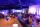 Une fête se déroule dans une grande salle. Les gens sont rassemblés autour de tables et se parlent entre eux en tenant des verres. La salle est éclairée de lumières violettes et bleues.
