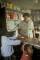 Une femme se tient derrière le comptoir d’un magasin. Deux jeunes enfants se tiennent devant elle. L’un fait face à la femme derrière le comptoir et l’autre regarde au loin.