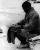 Une image en noir et blanc de Nelson Mandela. Il est assis et tient un vêtement sur ses genoux. Il semble être en train de coudre. Il porte des shorts, de longues chaussettes, des chaussures blanches et un veston.
