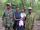 Une femme tenant un bébé, tandis que quatre hommes en tenue militaire se tiennent à côté et derrière elle. Ils sont tous debout devant une forêt et posent pour la photo.