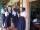 Un groupe de jeunes femmes et d’adolescentes debout sur la véranda d’un bâtiment. La plupart portent des blouses blanches et des jupes bleues, et deux filles portent aussi des chandails bleus. À droite, une porte mène vers une classe remplie d’élèves assises à des pupitres de bois et écrivant dans des carnets.