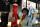 Une femme souriante se tient devant une robe de bal rouge et un smoking noir exposés sur des mannequins. La robe et le costume sont derrière une vitrine.