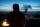 Au premier plan, la silhouette d’une personne qui semble courir vers nous, devant une voiture dont les phares sont allumés. À l’arrière-plan, un ciel bleu pâle avec la silhouette d’un arbre et des nuages longs et fins qui traversent l’horizon.