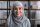 Une femme portant un hidjab bleu sourit vers l'objectif. Il y a une étagère de livres en arrière-plan.