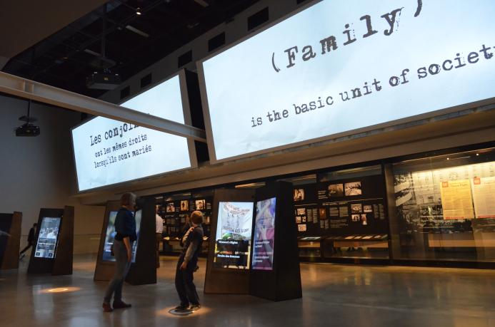 Une femme et un enfant interagissent avec un élément d'exposition numérique dans une galerie de musée. Au-dessus d'eux, sur un grand écran, on peut lire, en anglais, "La famille est l'unité de base de la société." Visibilité masquée.