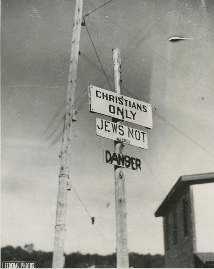 Black and white photo of three signs on a post, reading “Christians only,” “Jews not allowed” and “Danger.” Visibilité masquée.