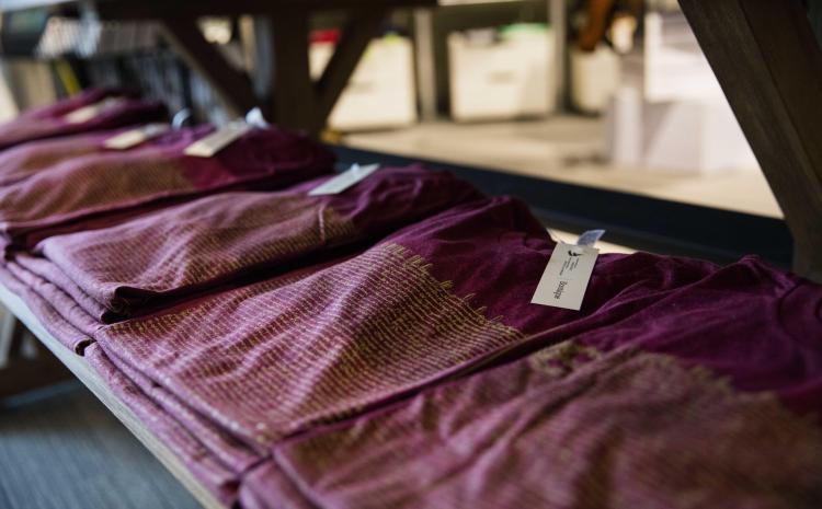 Des piles de trois ou quatre t-shirts couleur rouge vin pliés sont posées sur une table en bois. Visibilité masquée.