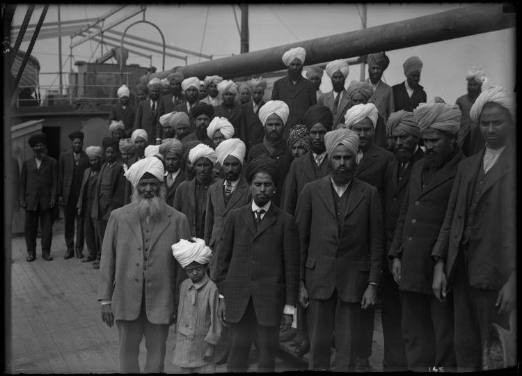 Une image en noir et blanc d’un grand groupe d’hommes sikh et d’un garçon debout sur le pont d’un navire. Ils font tous face au photographe et ne sourient pas.