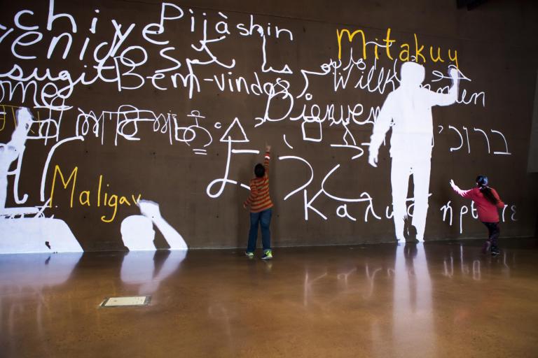A video of people’s silhouettes is projected onto a large wall. They are writing the word “welcome” in many languages. Two children interact with the video. Visibilité masquée.