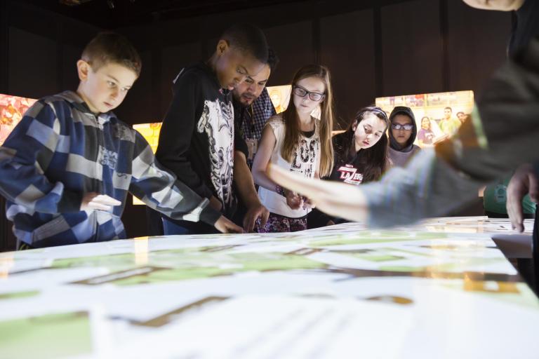 Un groupe d'élèves regardent la surface interactive d'une table qui émet une lumière blanche. Visibilité masquée.