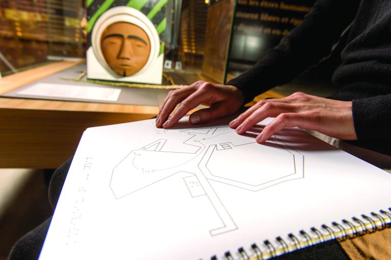 Les mains d'une personne lisent le braille sur une page d'un carnet à anneaux avec des motifs géométriques. Visibilité masquée.