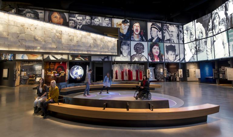 Cinq personnes dans une galerie du Musée. Deux personnes sont assises sur un banc. Trois personnes, dont l'une est en fauteuil roulant, sont dans une zone circulaire où des bulles de lumière colorée sont projetées sur le plancher. Visibilité masquée.