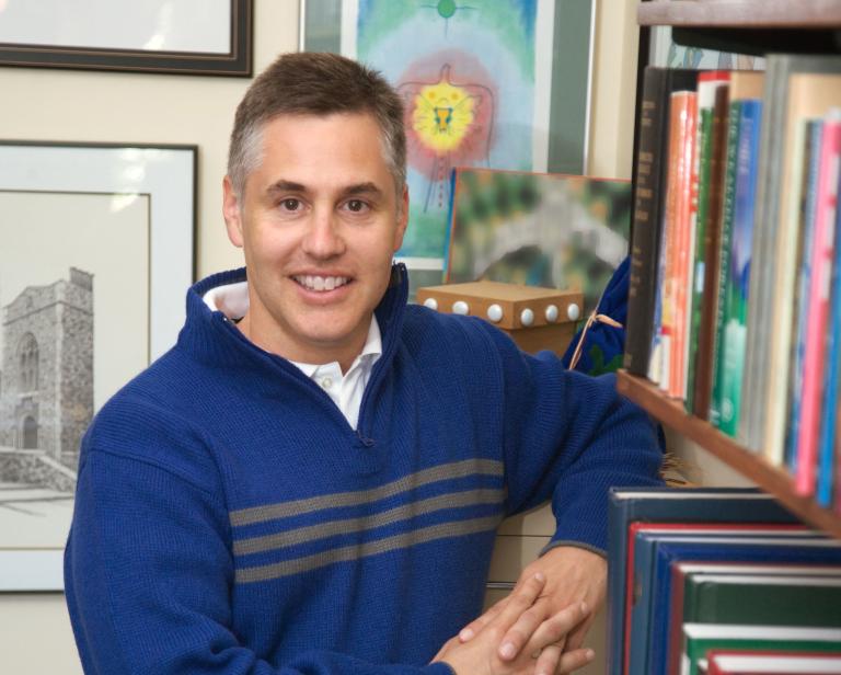 Un homme debout près d’une étagère de livres regard vers l’objectif. Visibilité masquée.