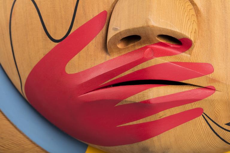 Un plan rapproché d’une boîte en bois, sur laquelle est sculpté un visage dont la bouche est recouverte d’une main peinte en rouge. Visibilité masquée.