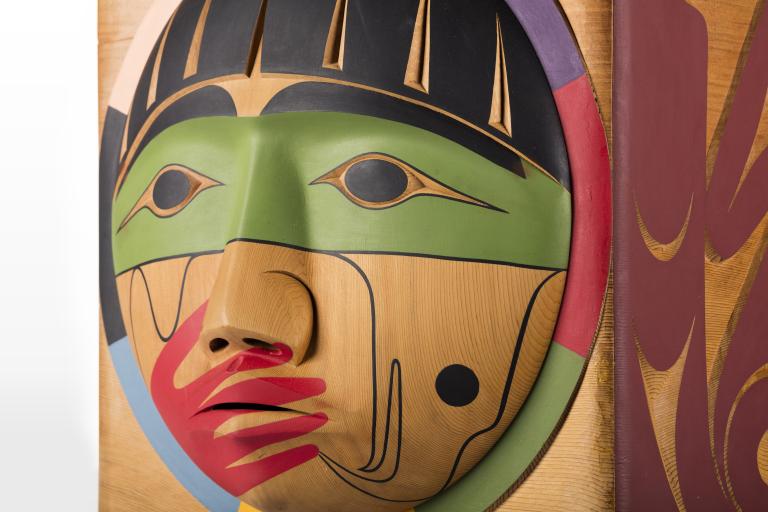 Un plan rapproché d’une boîte en bois, sur laquelle est sculpté un visage dont la bouche est recouverte d’une main peinte en rouge. Visibilité masquée.