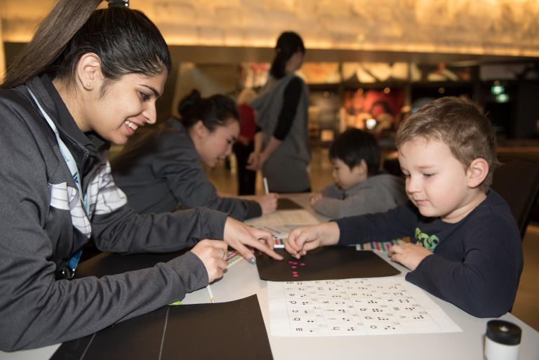 A Museum staff member helping a young child place coloured dots in the shape of braille letters. Visibilité masquée.