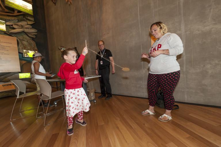 A young girl holds a small stick attached to a string and a small circular piece of cardboard. She is swinging this rope and cardboard as two adults watch. Visibilité masquée.